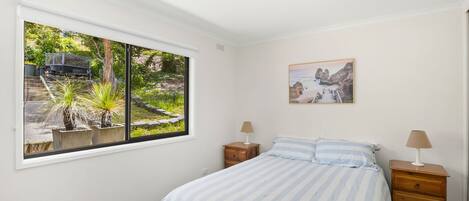 A bedroom with a bed, two bedside tables, a window with a view of a garden and a parked vehicle, and a painting on the wall.