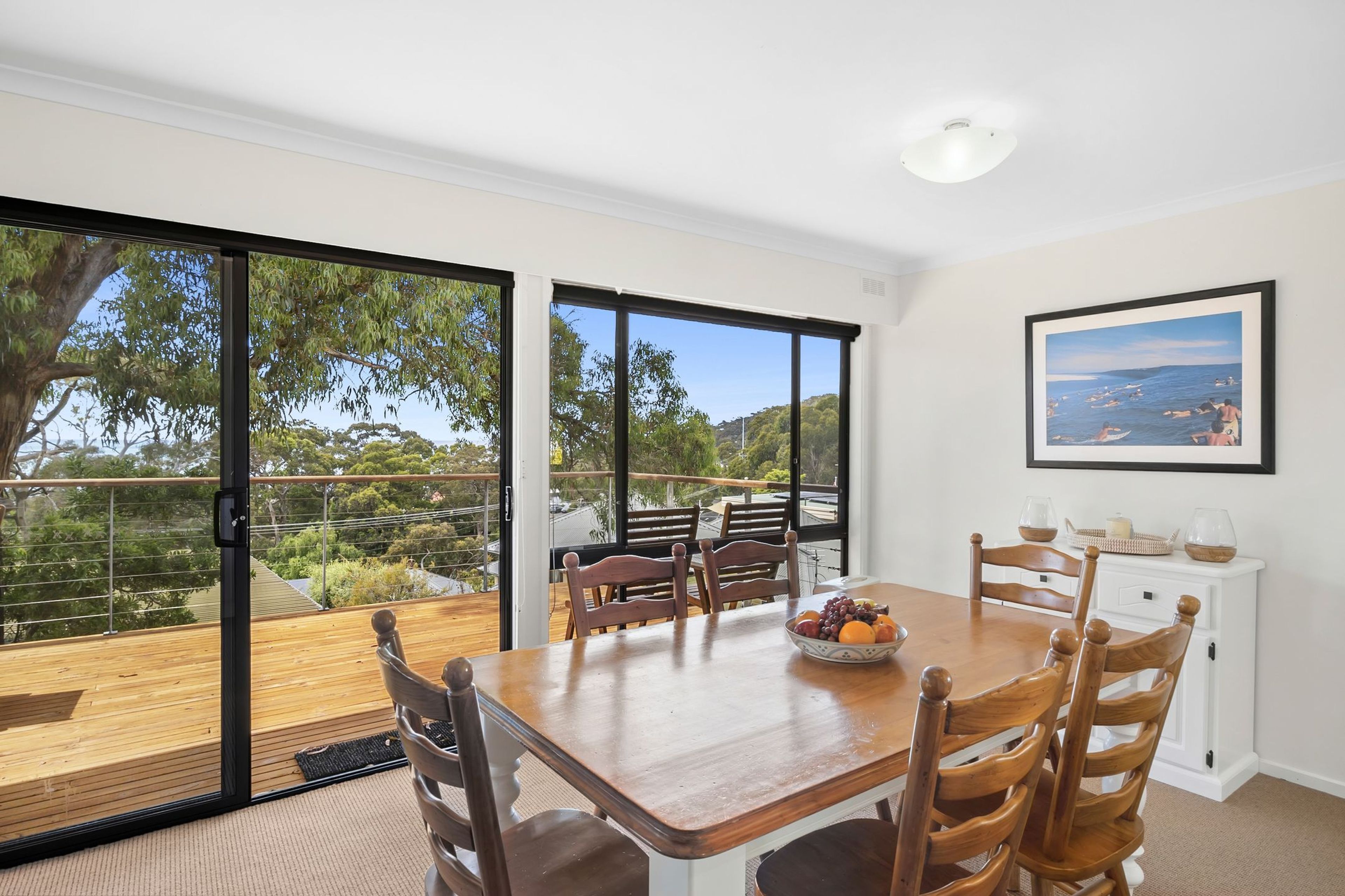 A dining area with a wooden table and chairs, a bowl of fruit, and a view of a coastal landscape through large glass doors.