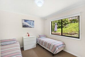 Een slaapkamer met twee eenpersoonsbedden, een witte dressoir, een plafondlamp, een raam met uitzicht op groen en een ingelijst kunstwerk aan de muur.