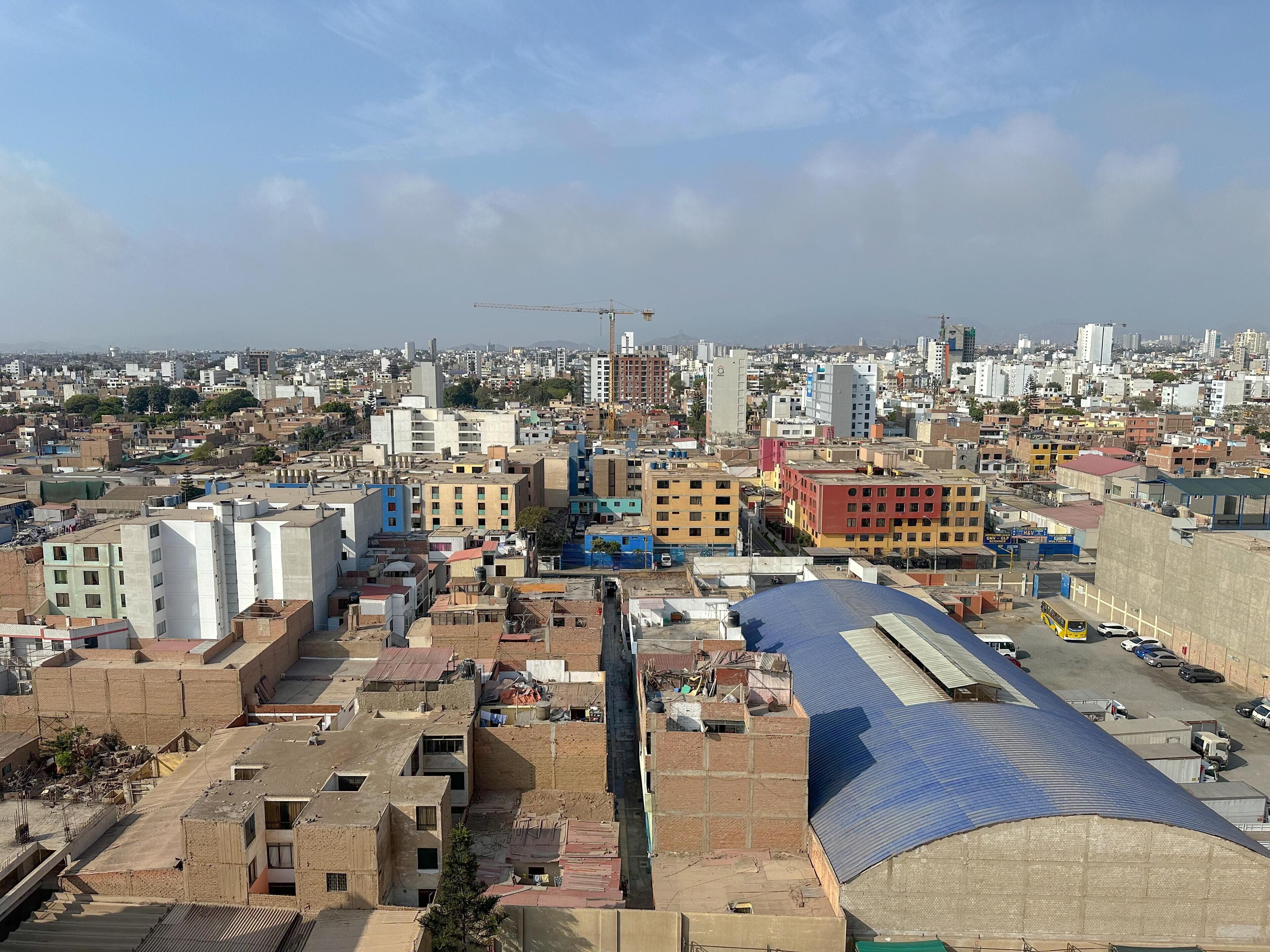 Un paisaje urbano con una combinación de edificios residenciales y comerciales, una estructura con techo azul destacada, y una grúa de construcción a lo lejos.