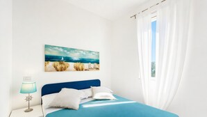 A bedroom with a bed, a nightstand with a lamp, a painting of a beach scene, and white curtains. - Apartment 'Le Case Di Chri' with Wi-Fi and Air Conditioning (Mattinata)