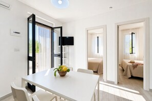 A modern hotel room with a white table, a flat-screen TV, a bed with a beige bedspread, and a window with sheer curtains. - Le Case Di Chri Apartment with Wi-Fi and Air Conditioning (Mattinata)