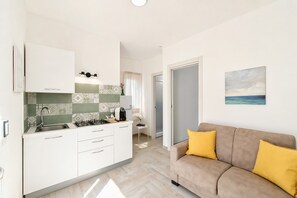 A modern kitchen with white cabinets, a gas stove, and a sink. There is a dining area with a white chair and a small table. A beige sofa with yellow cushions is in the living area, and a painting of a seascape is on the wall. - Apartment 'Le Case Di Chri' with Wi-Fi and Air Conditioning (Mattinata)