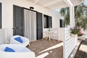 Un espace extérieur abrité avec des chaises blanches et une table, une porte avec des volets noirs et un pot de fleurs.