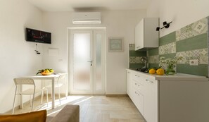 Una cocina con armarios blancos, una mesa de comedor con sillas, un televisor montado en la pared y una puerta que da a otra habitación.