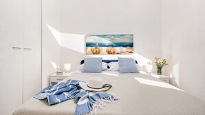 Un dormitorio con cama, almohadas, un sombrero, una bufanda, un cuadro, una mesita de noche, una lámpara y un florero con flores.