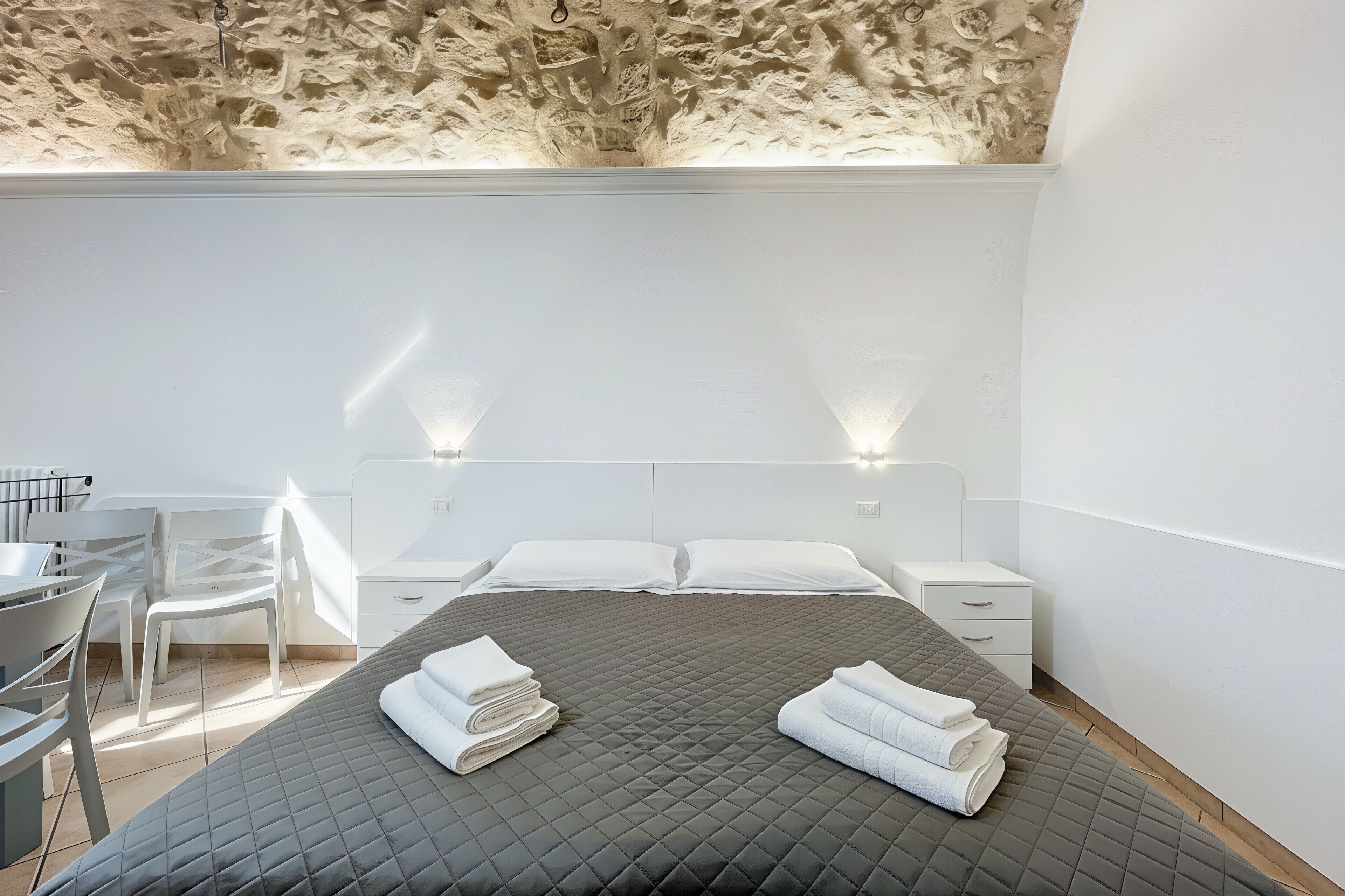 A modern hotel room with a bed, two bedside tables, a desk with chairs, and a ceiling with a unique texture.