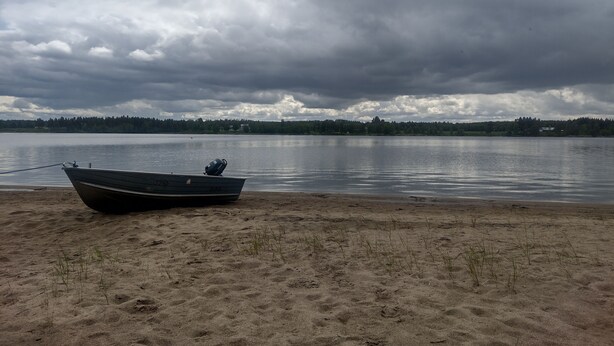 En båt på en sandstrand med en stilla sjö och en molnig himmel i bakgrunden.