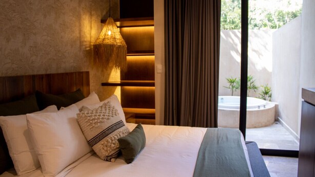 A modern hotel room with a large bed, a bedside lamp, a view of a garden and a hot tub outside. - Peregrina Tulum 102A 2BR Luxury Condo (Tulum)