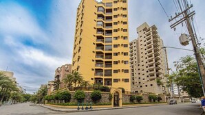 A tall yellow apartment building with multiple balconies, a stone entrance, and a paved street with parked scooters. - Apto com Garagem e Ar-condicionado a 600m da Praia (Guarujá)