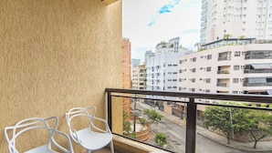 A balcony with two white chairs and a small table, overlooking a cityscape with buildings and a street. - Apto com Garagem e Ar-condicionado a 600m da Praia (Guarujá)