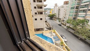 A view from a window overlooking a street with a pool area and a building with multiple floors. - Apto com Garagem e Ar-condicionado a 600m da Praia (Guarujá)