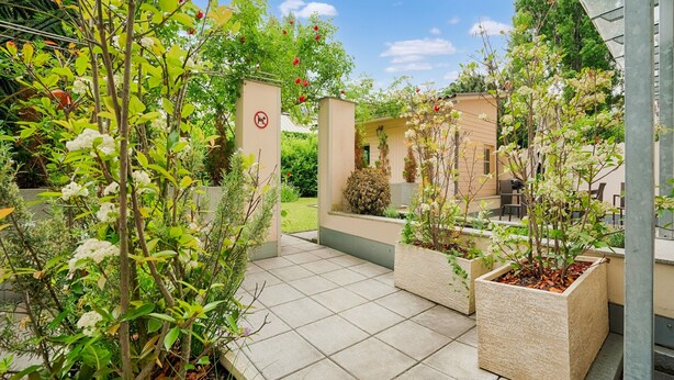 Un jardín con un sendero de baldosas, plantas en macetas y un edificio con fachada color crema.