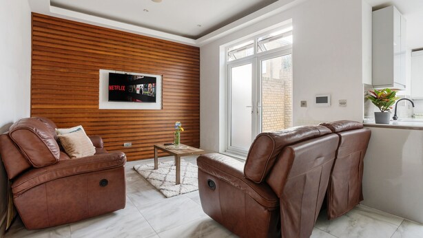 A modern living room with leather sofas, a wooden accent wall, a TV, and a large window. - 5 BR Flat |Spacious|Near Shepherd's Bush Stn.|ST (Greater London)