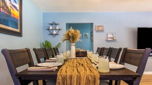 Una mesa de comedor con mantel rústico, rodeada de sillas de madera oscura, dispuesta con platos, vasos y un centro de mesa.