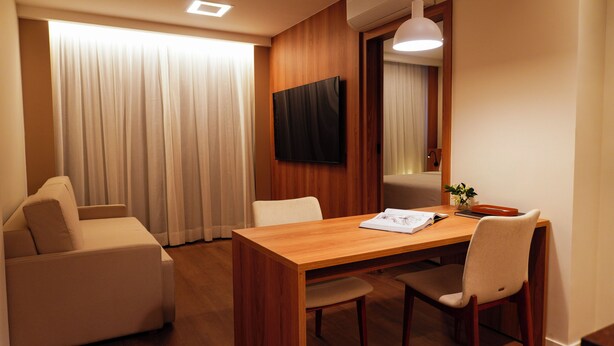 Habitación de hotel con una mesa de comedor de madera, dos sillas, un sofá, un televisor montado en la pared y un ventanal con cortinas.