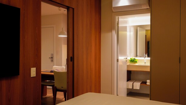 Habitació d'hotel amb una porta de fusta, un escriptori amb una cadira, un llit, un bany amb un mirall i una planta sobre el lavabo.