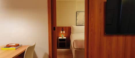 A hotel room with a wooden desk, a chair, a bed, a nightstand with a lamp, and a wall-mounted TV.