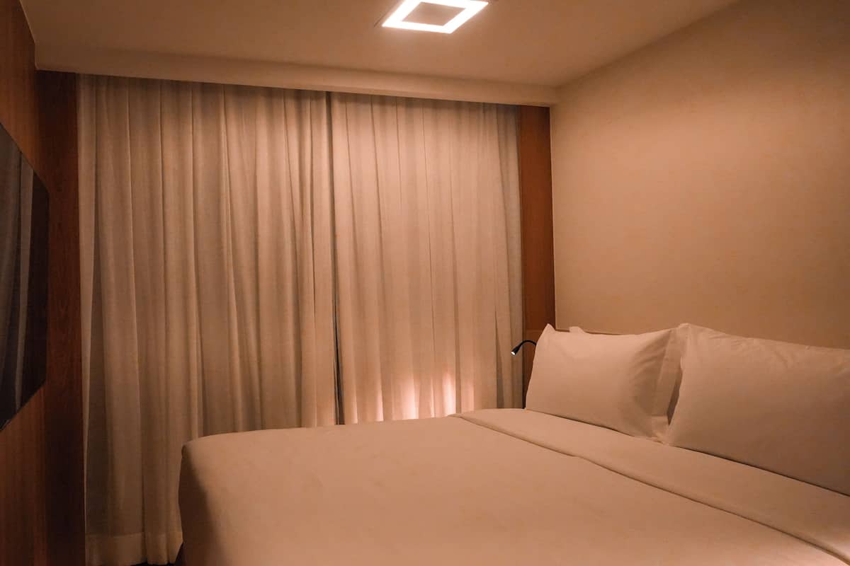 Habitación de hotel con cama, cortinas, televisor montado en la pared y una lámpara en el techo.