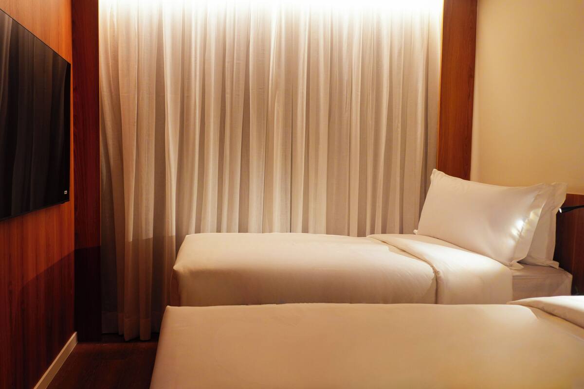Habitación de hotel con dos camas, un televisor montado en la pared y un amplio ventanal con cortinas.