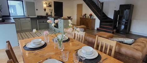 A dining area with a wooden table set for four, surrounded by leather sofas. A kitchen and living area are visible in the background.