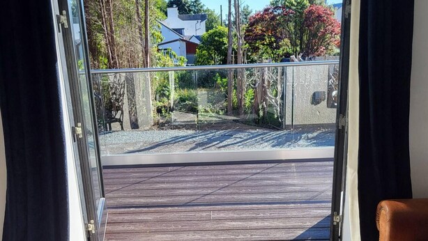 Udsigt fra en balkon med træterrasse og gel ænder af glas, samt et glimt af træer og huse i baggrunden.