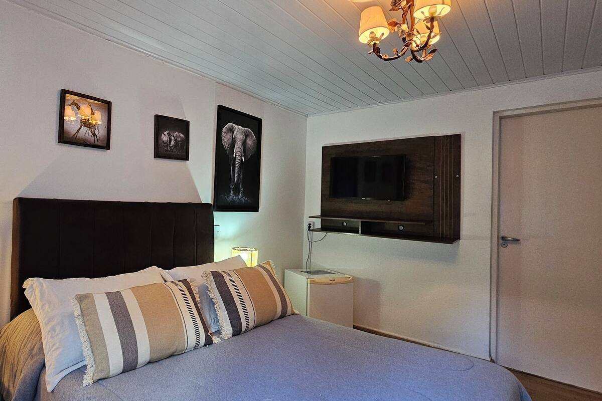 Ein Schlafzimmer mit Bett, Nachttisch mit Lampe, an der Wand montiertem Fernseher und gerahmten Bildern.
