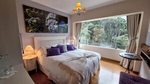 Quarto com uma cama grande, mesa de cabeceira, abajur, janela com cortinas e vista para árvores.