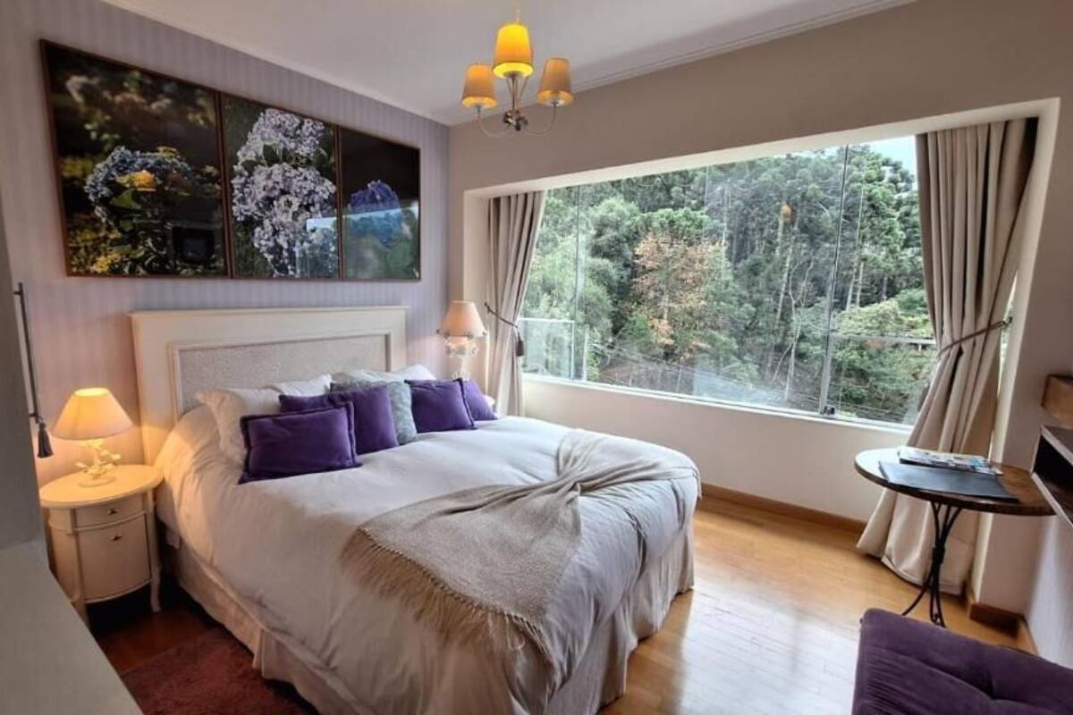 Ein Schlafzimmer mit einem großen Bett, einem Nachttisch, einer Lampe, einem Fenster mit Vorhängen und Blick auf Bäume.
