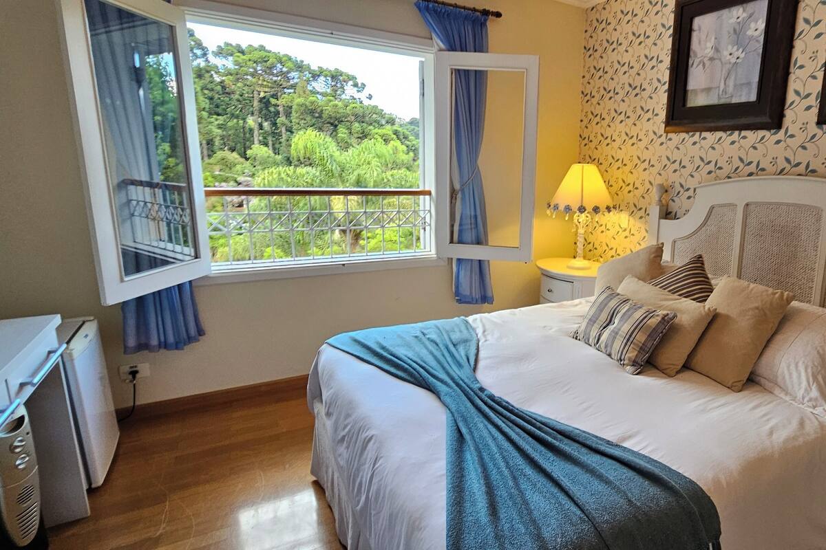 Ein Schlafzimmer mit Bett, Nachttisch, Lampe, Fenster mit Vorhängen und Blick auf Grünfläche.