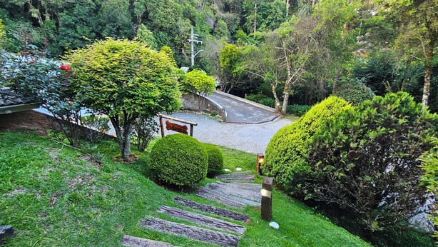 Uma trilha leva a um edifício com telhado tradicional, cercado por vegetação exuberante e um jardim bem cuidado.