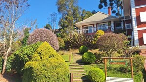 Um jardim bem cuidado, com arbustos aparados e um caminho que leva a uma casa com fachada vermelha e telhado de telhas.