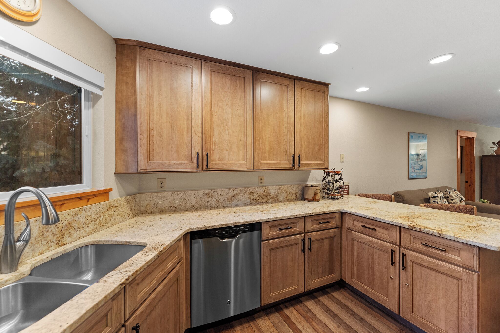 A kitchen with wooden cabinets, a stainless steel dishwasher, granite countertops, and a window with a view of trees.