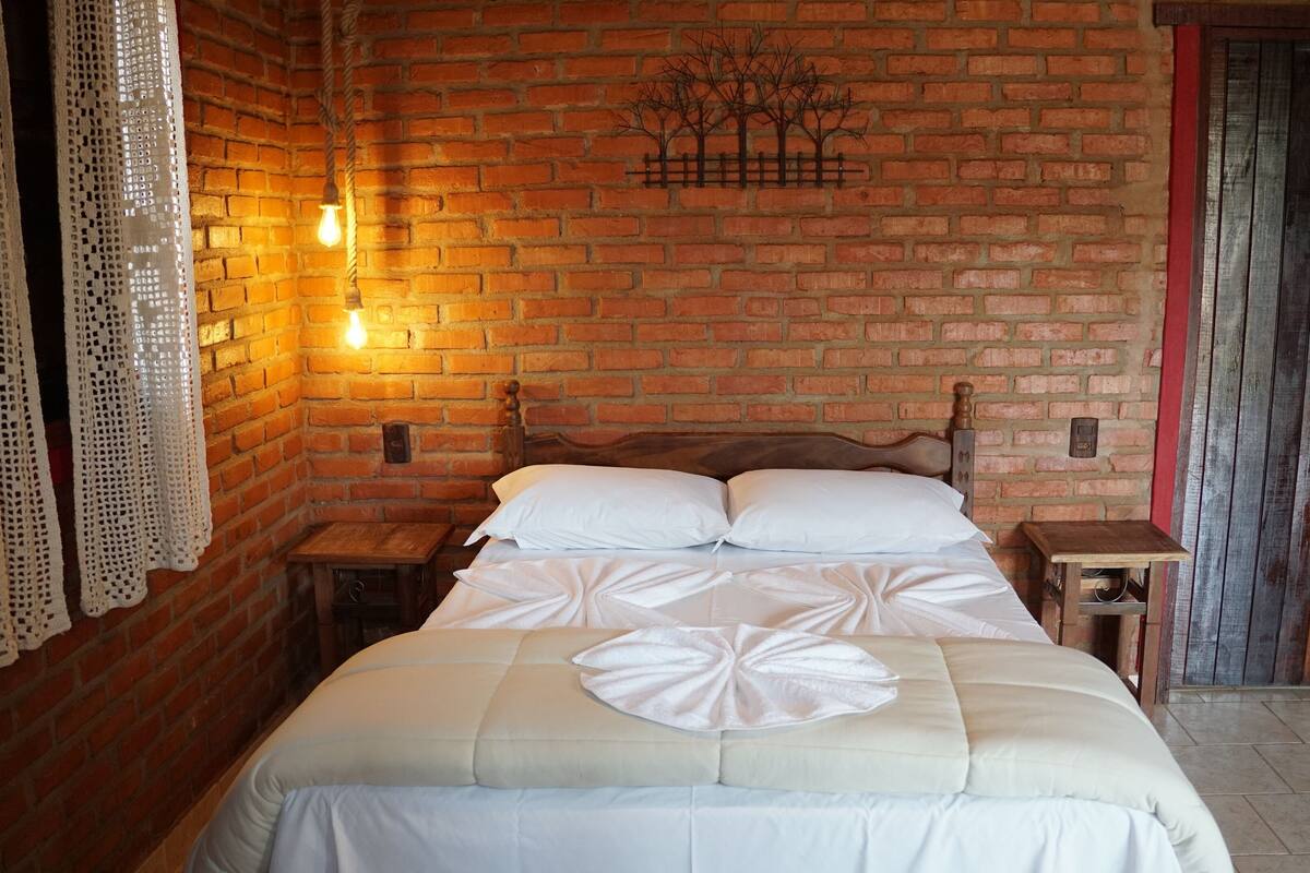 A room with a brick wall, a bed with white bedding, a wooden nightstand, and a wall-mounted light fixture.