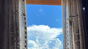 A view through a window with a wooden frame and lace curtains, overlooking a green hillside and a partly cloudy sky.