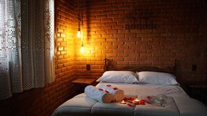 A cozy room with a brick wall, a bed with white linens, a wooden headboard, a nightstand, and a lit wall sconce.