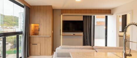 A modern hotel room with a large window, a built-in TV, a sofa, a small kitchen area, and a bathroom with a modern sink and mirror.