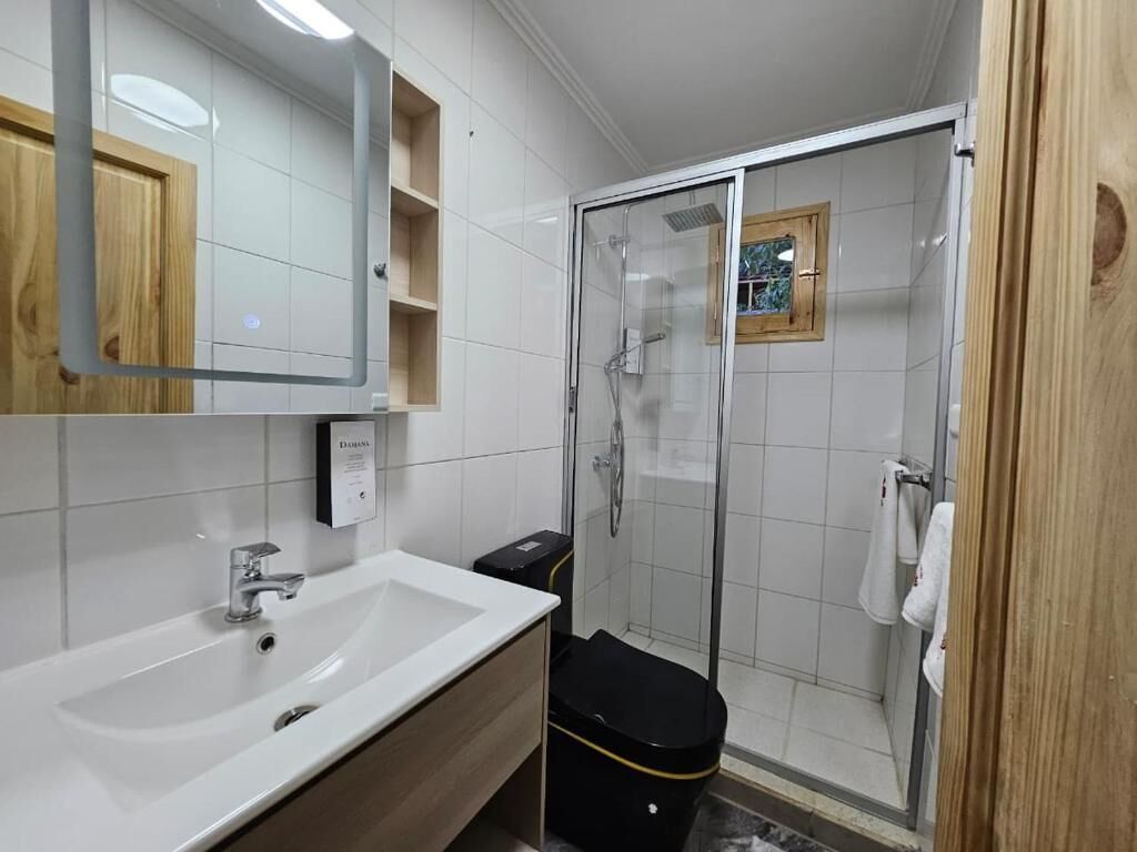 Un baño con un lavabo blanco, un espejo, una ducha con cerramiento de vidrio y un cubo de basura negro.
