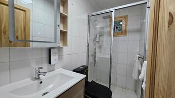 Un baño con un lavabo blanco, un espejo, una ducha con cerramiento de vidrio y un cubo de basura negro.