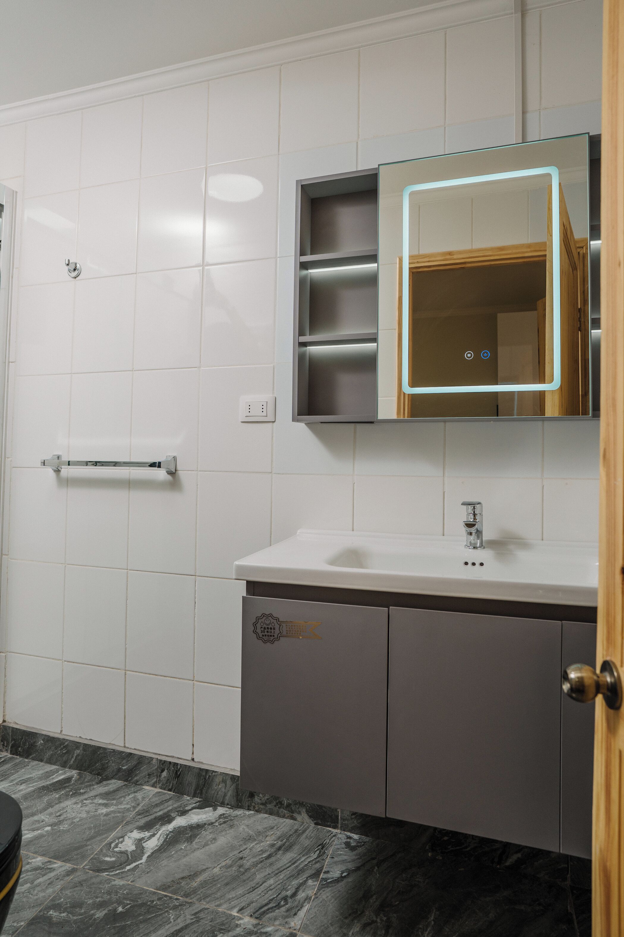 Un baño con paredes de azulejos blancos, suelo de mármol, lavabo blanco, un espejo con luz y un mueble de madera.