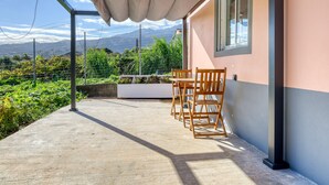Zona salotto all'aperto riparata, con tavolo e sedie, pavimento in cemento e vista su una catena montuosa attraverso una recinzione metallica.