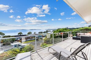 A deck with outdoor furniture overlooking a scenic view of the sea and a residential area. - Panui Views - Snells Beach Holiday Home (Snells Beach)