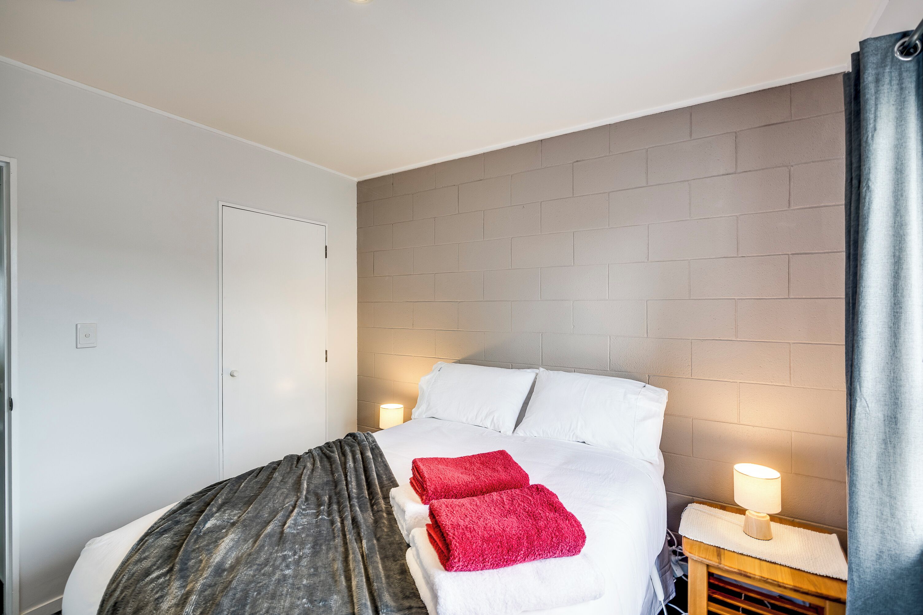 Ein ordentlich bezogenes Bett mit weißen Leinen und zwei roten Handtüchern, eine Nachttischlampe und ein Nachttisch aus Holz.