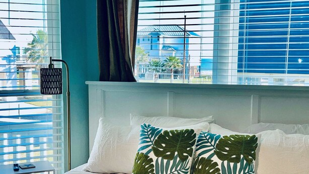 A hotel room with a bed, white bedding, and a view of a blue building through large windows with blinds. - Saltwater Solace, For 8 (Galveston)
