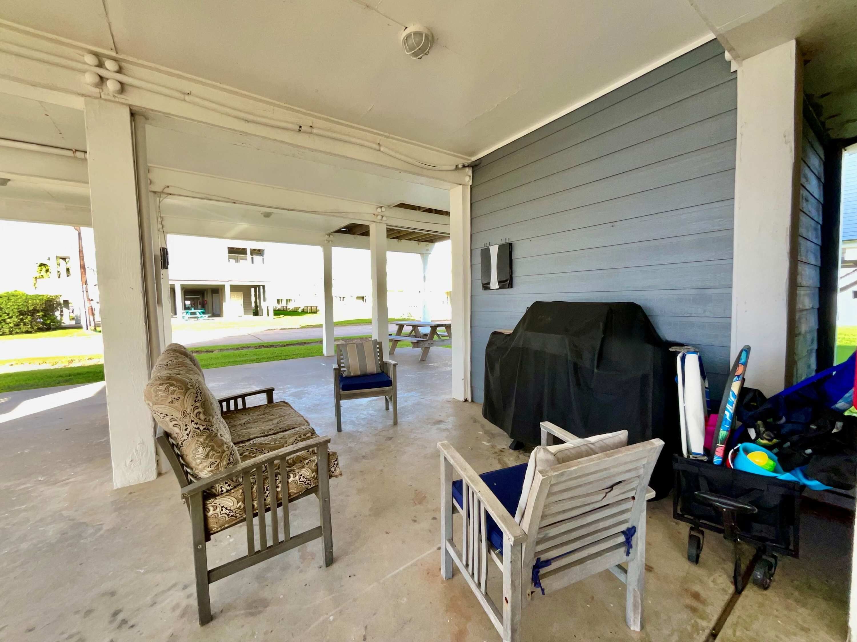 A covered outdoor area with a patio chair, a cushioned chair, a grill, and a stroller.
