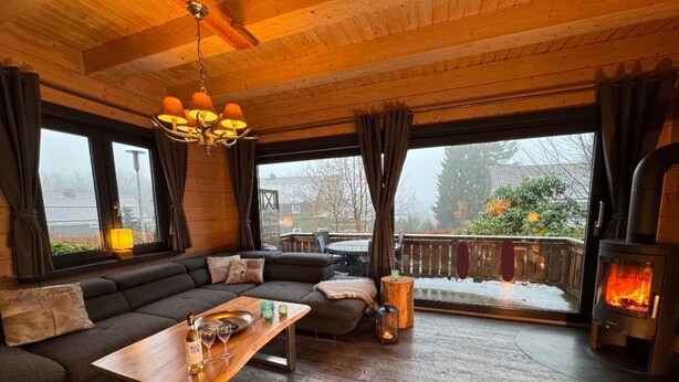 A cozy room with a wooden ceiling, a sofa, a table with glasses and a bottle, a fireplace, and large windows. - Blockhutte im Harz mit Sauna und Kamin (Goslar)