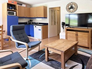 Un espace de vie compact comprenant un coin cuisine, un coin repas avec une table et des chaises, un téléviseur et une horloge murale.
