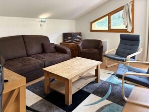 Ein möbliertes Zimmer mit einer Couch, zwei Stühlen, einem Holztisch, einem Teppich und einem Fenster mit Vorhängen.