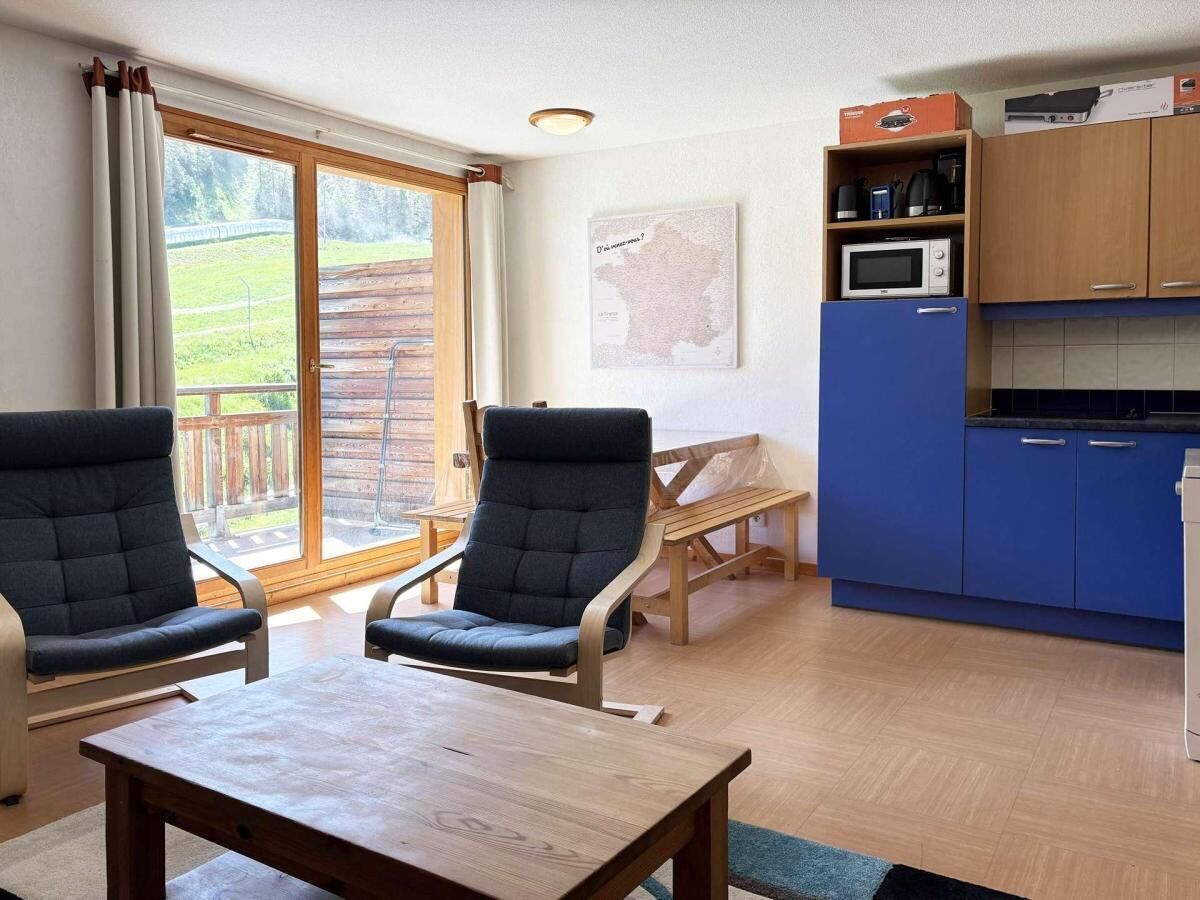 Ein Zimmer mit zwei Stühlen, einem Tisch, einer Küchenzeile mit blauen Schränken und Blick auf eine Holzterrasse durch ein großes Fenster.