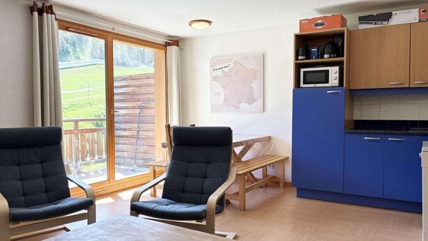 Ein Zimmer mit zwei Stühlen, einem Tisch, einer Küchenzeile mit blauen Schränken und Blick auf eine Holzterrasse durch ein großes Fenster.